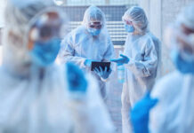 Healthcare workers in protective gear discussing information on a tablet outdoors