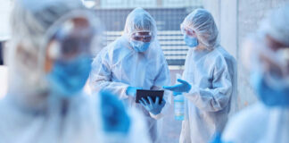 Healthcare workers in protective gear discussing information on a tablet outdoors