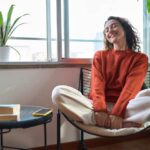 A relaxed woman sitting in a chair with a smile, surrounded by plants and natural light