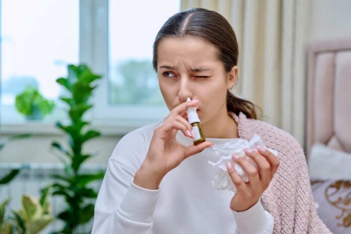 Woman applying nasal spray while holding a tissue in a cozy indoor environment